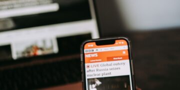 Person reading breaking news on a smartphone with a laptop in the background